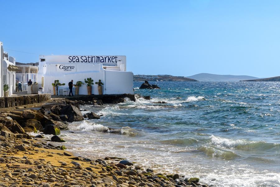 A low whitewashed building on a rocky shore with SEA SATIN MARKET painted large on its wall, cactus pots along the terrace, waves breaking over rocks in the foreground and an island on the horizon.