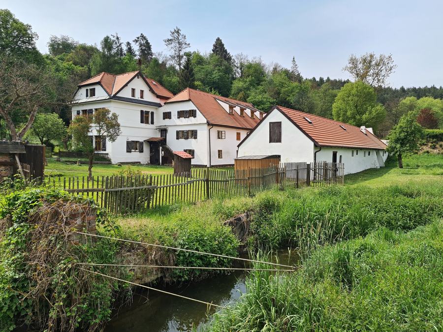 A white farmhouse with steep red roofs behind a wooden paling fence, a meadow in front, a narrow stream in the foreground with grass on both banks, wooded hills behind.