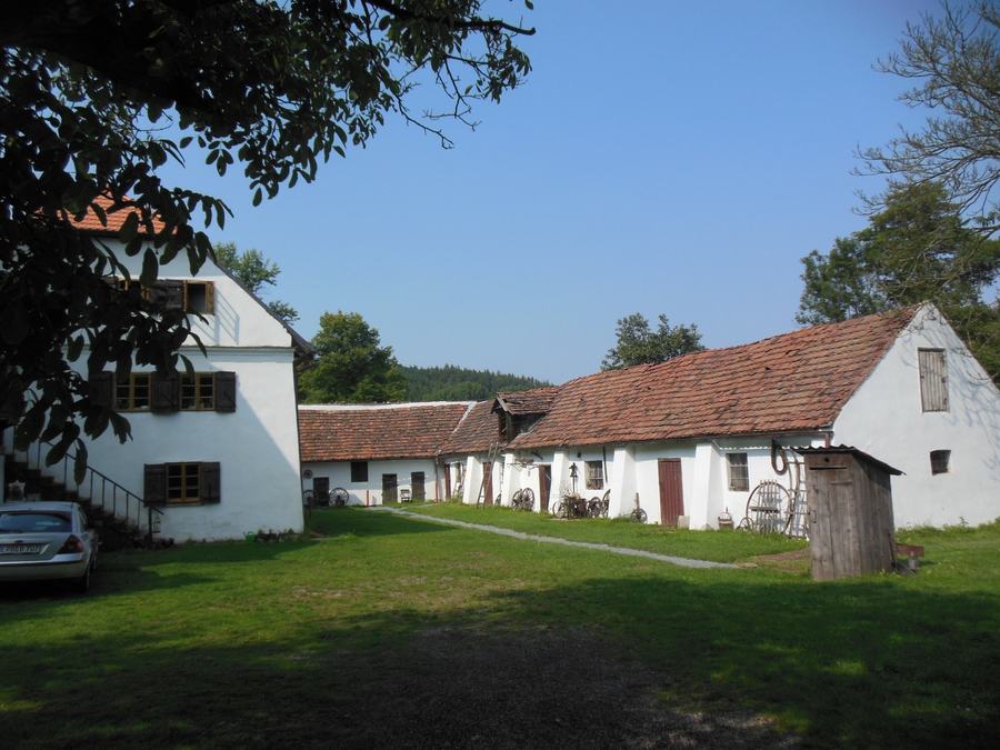 A whitewashed farmstead around a grassy yard, red tiled roofs, an outdoor wooden staircase on the left, old farm implements leaning along the wall of a long low barn, woods behind.
