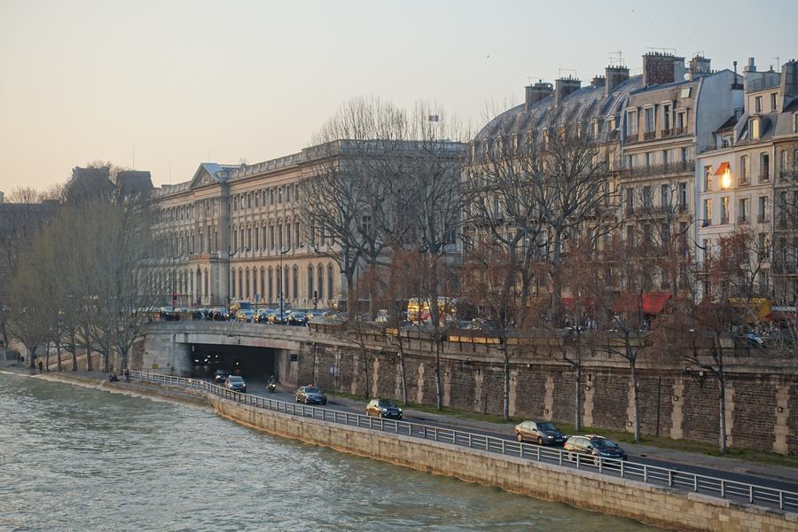 A riverside road running below a stone quay wall along the Seine, several cars on it, a tunnel entrance to the left, Haussmann buildings and bare winter trees above the wall in evening light.