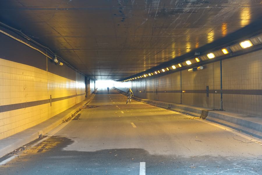 The inside of a wide road tunnel with tiled walls and rows of ceiling lights, no vehicles, a single rollerblader in the middle distance and daylight at the far end.