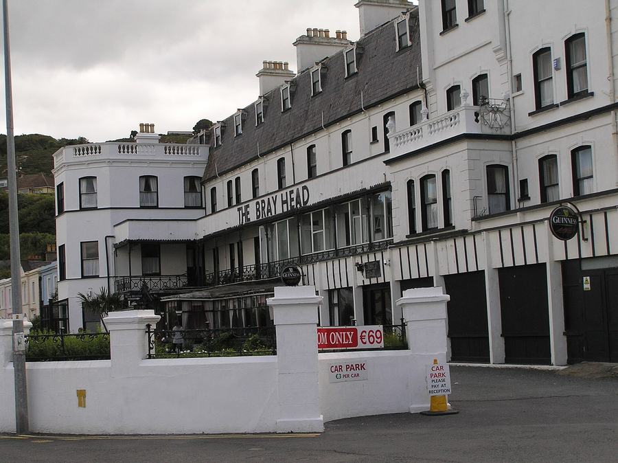 A large white Victorian seafront hotel with a slate mansard roof, dormer windows, tall chimney stacks and balustraded parapets, its name THE BRAY HEAD in dark letters along a first-floor canopy, Guinness signs by the entrance, a white boundary wall with car park notices in front and a wooded headland rising behind.