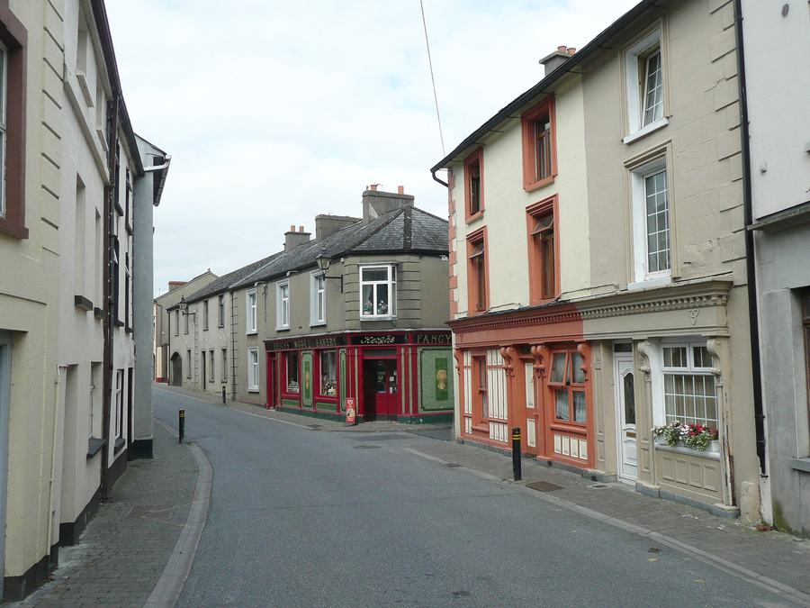 An empty curving street of two- and three-storey houses in Callan, rendered in cream, grey and terracotta, with sash windows and a decorated shopfront on the right; at the far corner a red and green tiled shopfront lettered KEOGH'S MODEL BAKERY.