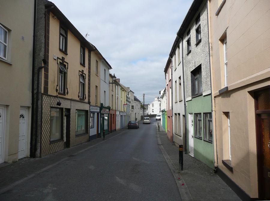 A narrow street of joined houses in pale yellow, pink, green and grey render, several with old shopfronts and empty display windows, one faced in patterned buff tiles; a few cars parked along the left kerb and the road rising to open sky at the far end.