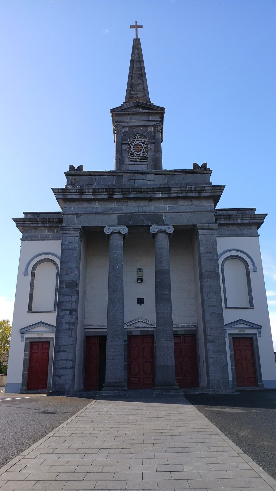 The west front of a grey limestone church seen straight on from the paved path: a pedimented portico on four tall Ionic columns, a red-painted double door in each of the five bays, plain white rendered wings either side with round-headed recesses, and above the roofline a square stone bell stage with a six-pointed star grille carrying a slender spire and a cross.