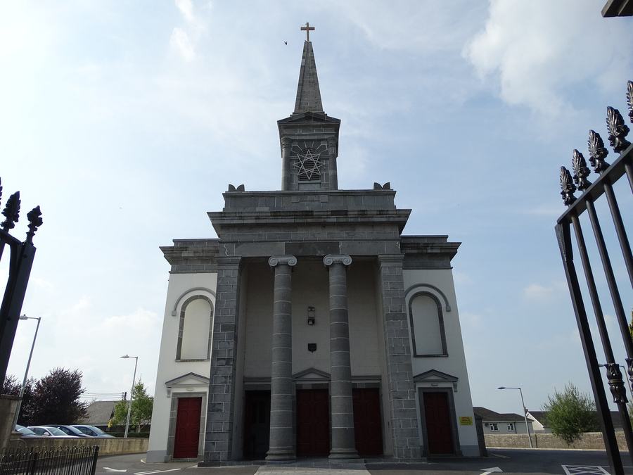 The same church front photographed from outside its railings, with an open wrought-iron gate leaf and spear-topped railings filling the right of the frame, parked cars and low houses to the left, and a wide tarmac forecourt in front of the portico.