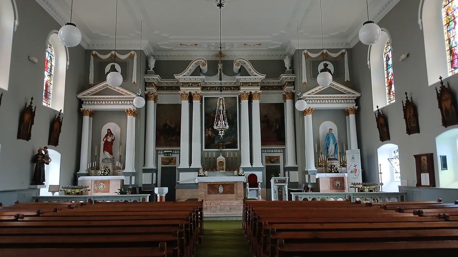 The interior of the church looking down the nave to the sanctuary: grey walls, white Corinthian pilasters and a broken scrolled pediment over a large painting of the Assumption, side altars with statues of the Sacred Heart and the Virgin under smaller pediments, stained glass in round-headed windows, white globe pendant lights, and rows of empty pine benches on a green carpet.
