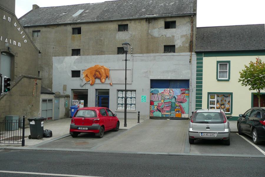 A stretch of Green Street in Callan: a tall bare-rendered three-storey warehouse with small unglazed window openings, a low grey frontage below it painted with a giant orange hand, a bright abstract mural filling a garage opening, an Irish-language sign on the gable to the left, a black lamp standard, and three parked cars along the kerb.