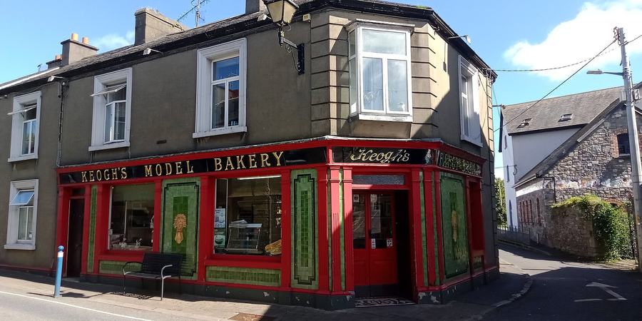 A corner shop with a scarlet-painted timber shopfront and glossy green tiled pilasters, gold lettering reading KEOGH'S MODEL BAKERY along the fascia and KEOGH'S FANCY BREAD around the corner; bread and a display cabinet visible through the plate glass, a bench outside, and a bay window on the first floor above.