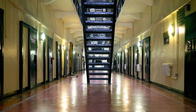 The ground floor of a prison wing seen down its length: red tiled floor reflecting the lamps, heavy dark cell doors with observation flaps in rows on both sides, and at the centre a black iron staircase and railed landings climbing three storeys towards a lit end wall.