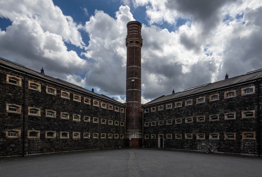 Two long three-storey wings of black basalt prison building meeting at an angle around an empty tarmac yard, each face gridded with small barred windows in pale stone surrounds, and between them a tall banded red-brick chimney rising into a sky of heavy white cloud.