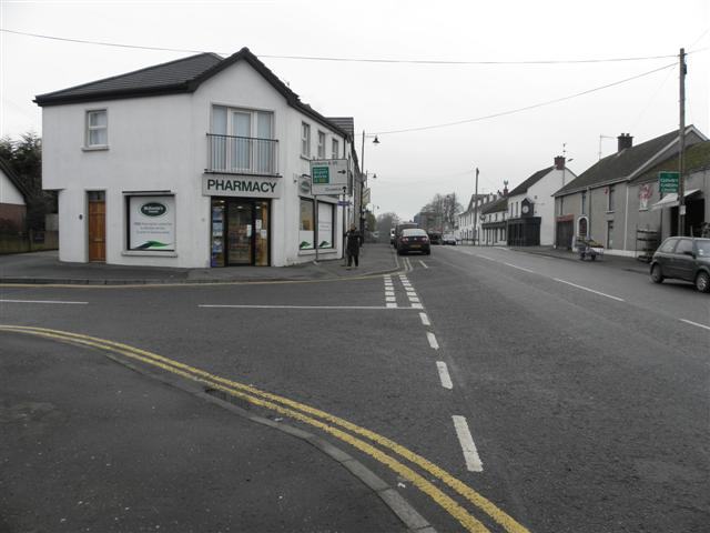 A wide empty road through a small village on an overcast winter day: a white two-storey corner building with a balcony and a black PHARMACY fascia on the left, a terrace of low houses and a few parked cars on the right, double yellow lines along the kerb and bare trees behind the roofs.