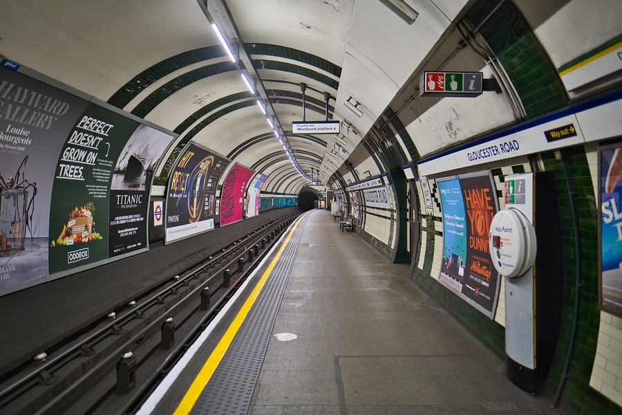 A deep-level tube platform curving away under a white tiled tunnel arch, advertisement panels along the far wall above the track, a yellow line on the platform edge, green and cream tiling on the right and a roundel-style nameboard reading GLOUCESTER ROAD, with an overhead sign for Piccadilly line Westbound platform 4.