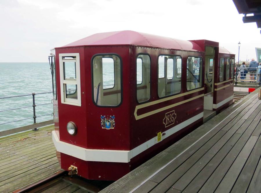 A short maroon and cream railway car standing on narrow-gauge track laid in the timber deck of a pier, the sea on one side and railings on the other, with the number 1835 and a coat of arms on its side and a round headlamp below.