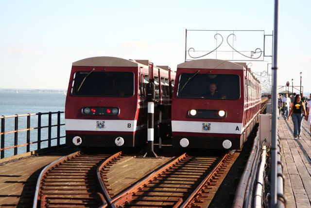 Two maroon and cream pier trains, lettered A and B, passing each other at a track loop on the open deck of Southend Pier, sea and hazy coastline to the left, a driver visible in the cab of train A and walkers on the boards to the right.