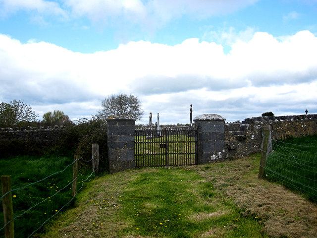 A plain iron gate between two squared limestone piers in a low field wall, seen along a grass track between wire fences; headstones and a Celtic cross show over the wall against a sky of broken cloud.