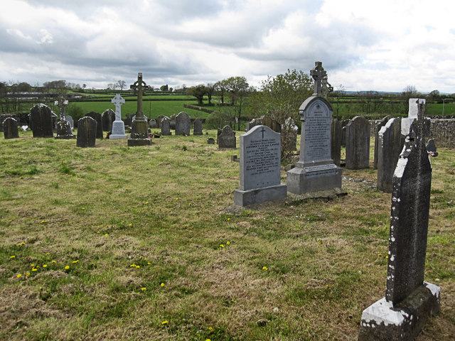 A small rough-grass burial ground on rising ground with scattered upright headstones, two tall Celtic crosses, a pair of polished granite memorials in the middle distance, dandelions in the grass, and green fields and hedgerows running away to a flat horizon beyond the boundary wall.