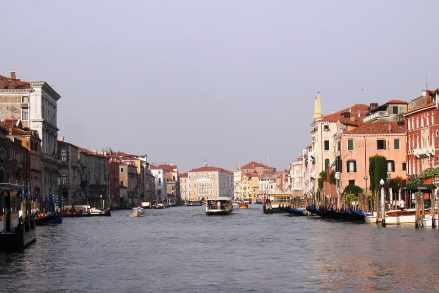 A wide canal lined on both sides with weathered Venetian palaces, a water bus in midstream and moored gondolas under blue covers along the right-hand quay.
