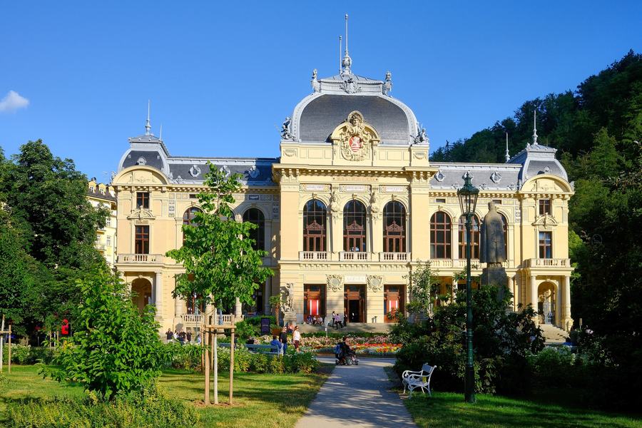 A pale yellow neo-Renaissance spa building with a grey mansard roof and a coat of arms over the central bay, seen across a garden with flowerbeds, young trees and a white bench; wooded hills rise behind it.
