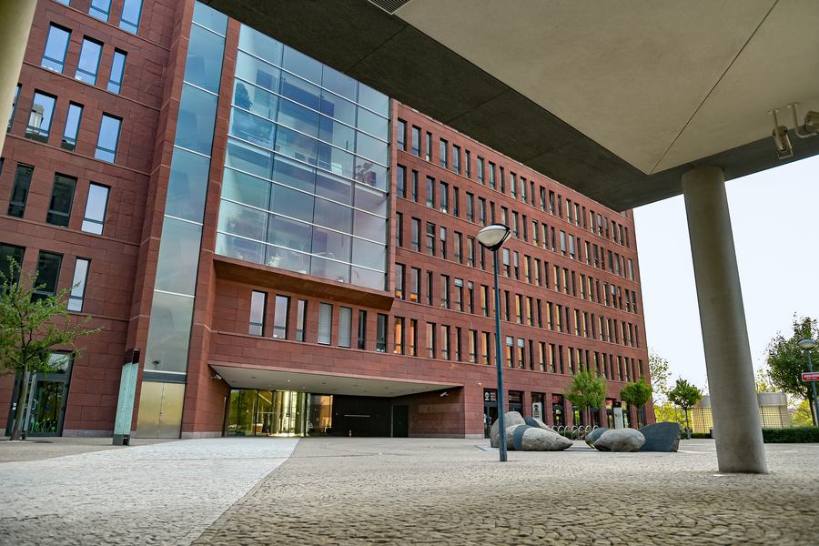 A modern red-brick and glass office block of six storeys with a recessed entrance, seen from a paved plaza with young trees and stone boulders, under the concrete soffit of a neighbouring building.