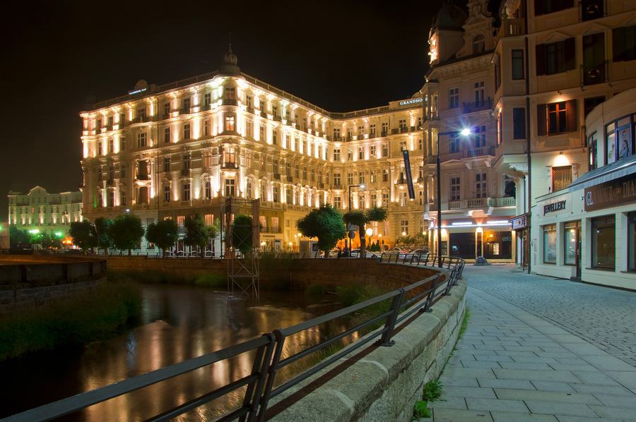 A large floodlit nineteenth-century hotel of five storeys reflected in a narrow river at night, with an empty paved embankment and railing in the foreground.