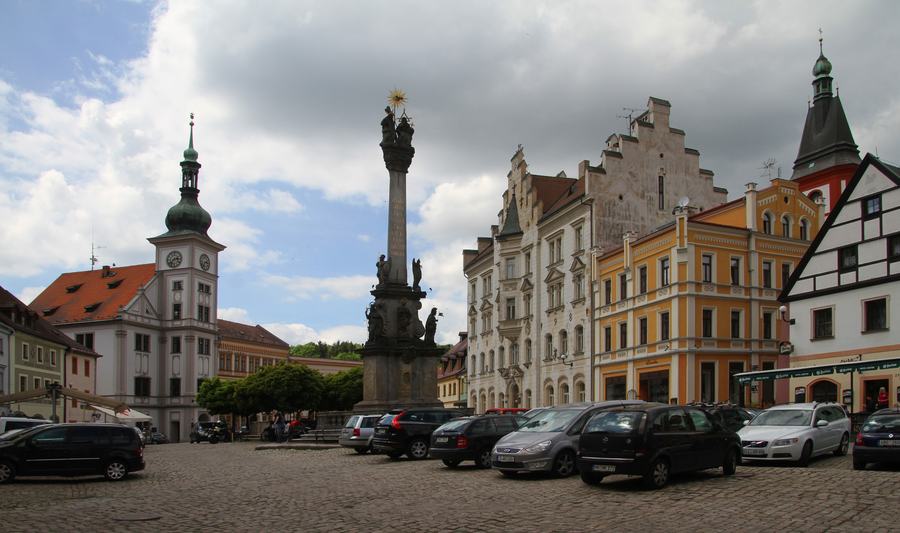 A cobbled town square ringed by gabled houses in white, ochre and green, with a baroque plague column at its centre, a church tower to the left and cars parked along the near side.
