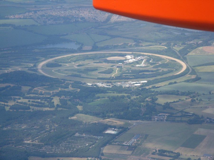An aerial view of a vehicle proving ground: a large oval banked test track enclosing winding roads and low buildings, surrounded by green Bedfordshire fields.