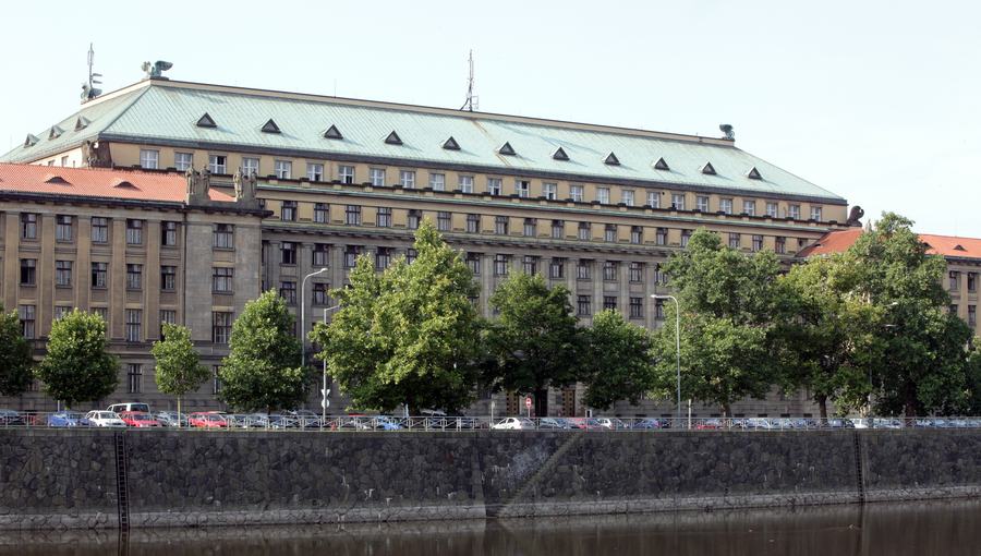 A long neoclassical government building with a green copper roof seen from the far bank of a river, a line of trees and parked cars along the quay in front of it.