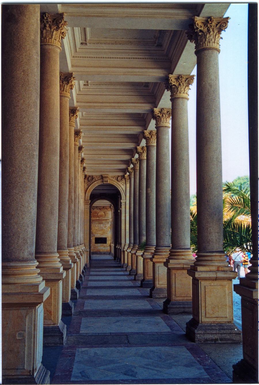 A long covered walkway receding into the distance between two rows of tall Corinthian columns, with a stone-flagged floor and a coffered ceiling.
