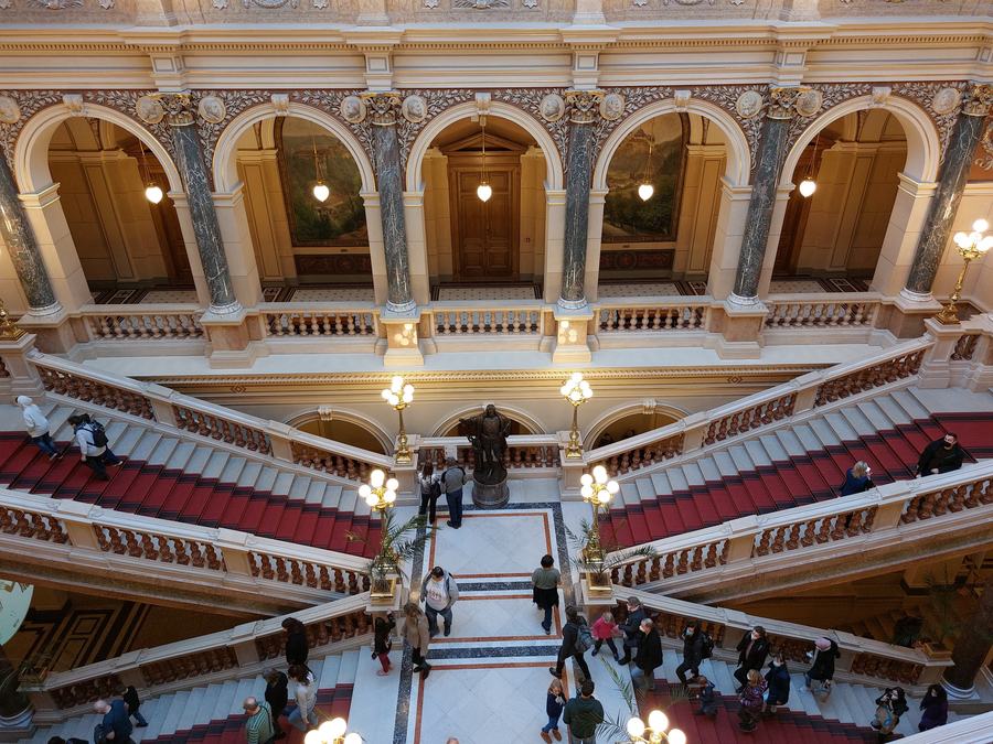 A grand nineteenth-century museum interior seen from above: twin marble staircases with red carpet running up either side of a bronze statue, arcades and painted lunettes behind, and visitors crossing the patterned stone floor.