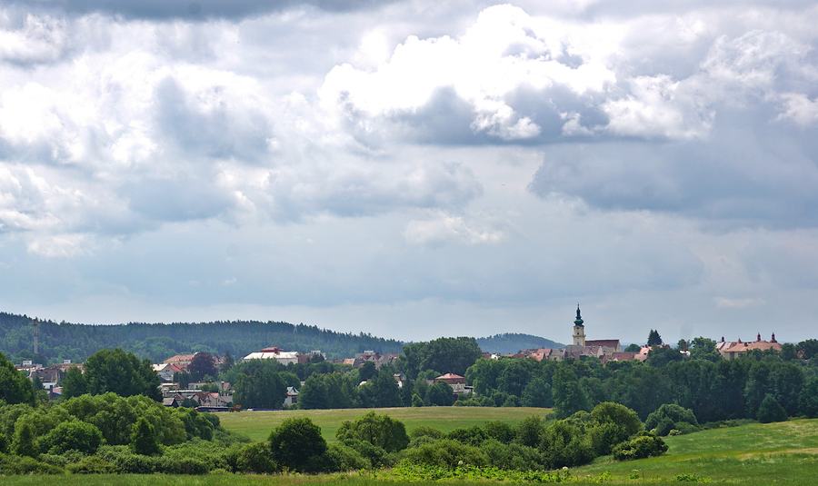 A small Czech town in the middle distance under a heavy cloudy sky, its church tower rising above low roofs, with meadows and lines of trees in the foreground.
