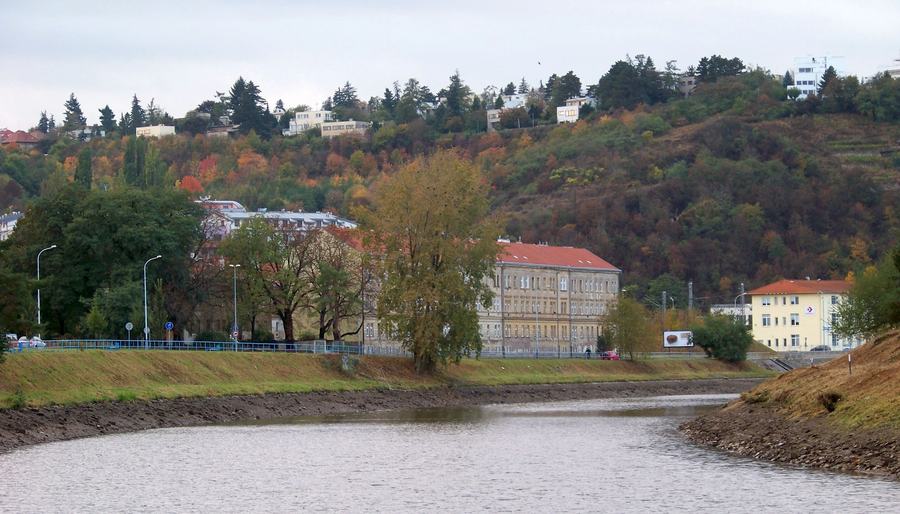 A river bend in autumn with a low weir, a road and a long yellow institutional building on the far bank, and a wooded hillside dotted with villas rising behind.