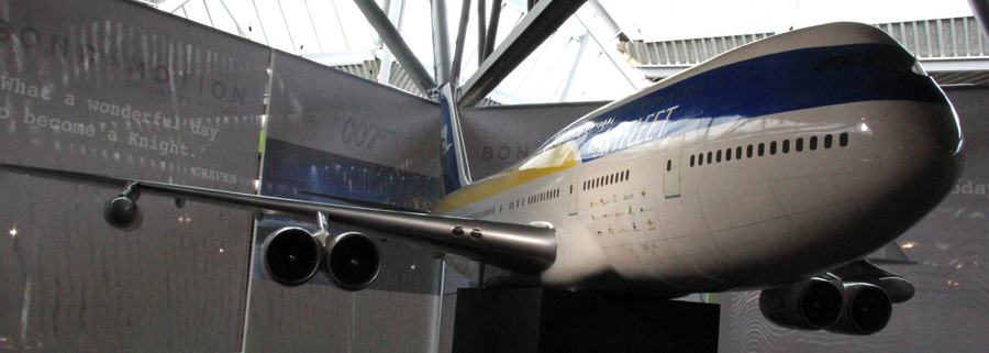 A large model airliner with a white and dark blue fuselage suspended in an exhibition hall, its nose pointing to the right, with lettering on the wall behind.