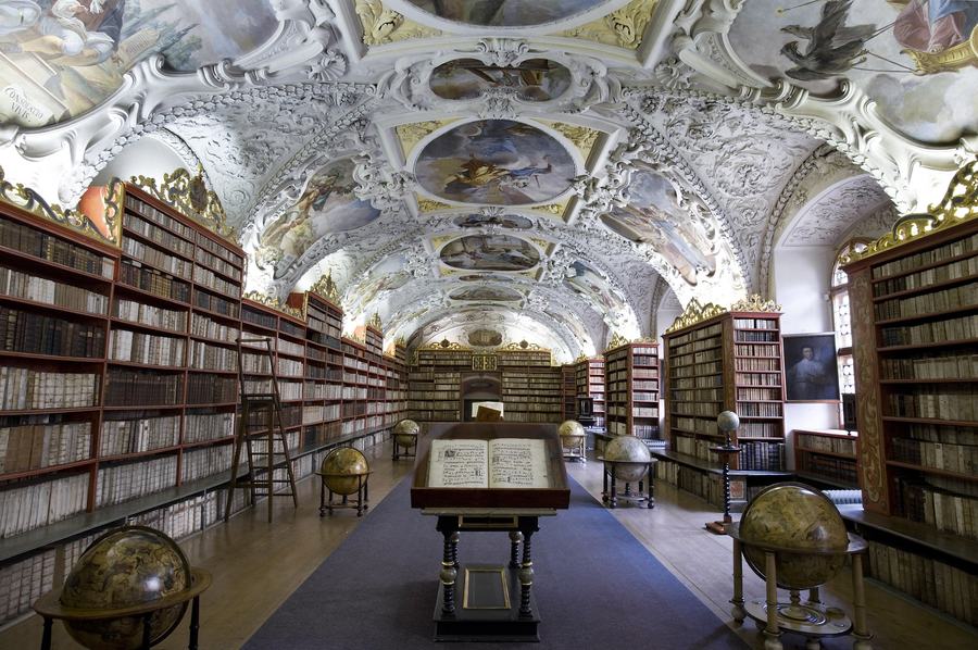 A barrel-vaulted baroque library hall with white stucco framing painted ceiling panels, walls of shelved leather-bound books, wooden globes on stands along the floor and an open folio on a table in the centre.