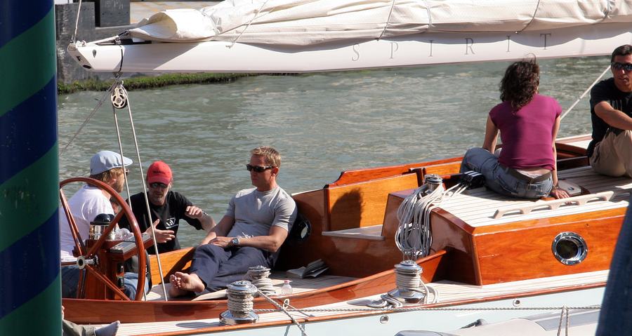 Several people sitting in the open cockpit of a varnished wooden sailing yacht moored in a canal, one of them in a grey T-shirt and sunglasses; the word SPIRIT is printed on the furled sail above them.
