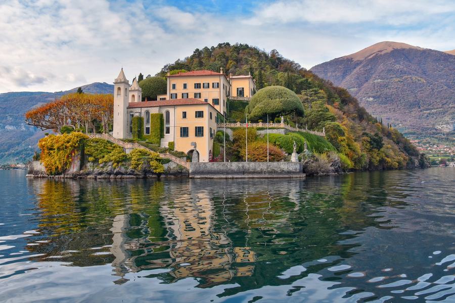 A pale ochre villa with a bell tower on a wooded promontory dropping straight into a calm lake, its terraced gardens and clipped trees reflected in the water, mountains beyond.