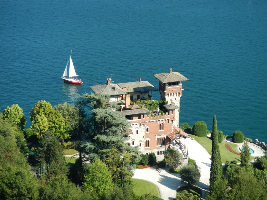 An aerial view of a brick and stone castellated villa with a square tower on a wooded promontory beside a deep blue lake, its lawns and drive running down to the shore, with a white sailing yacht on the water beyond.