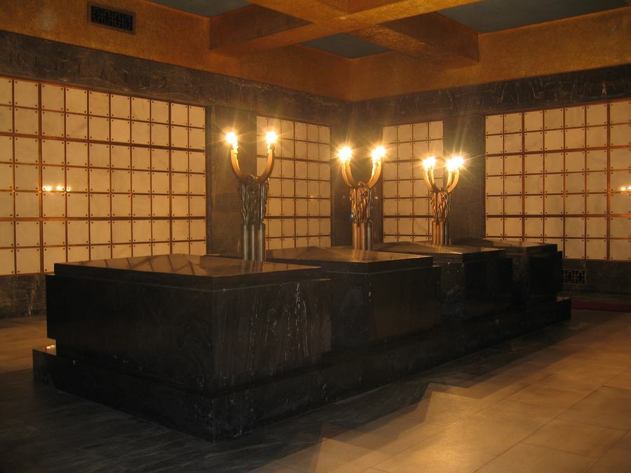 A dark marble funerary hall with three black sarcophagi in a row, each with a lit bronze candelabrum behind it, and walls of small square columbarium plaques.