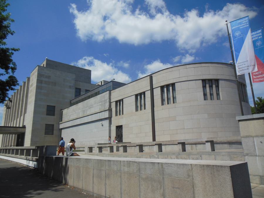 A severe granite-faced functionalist monument with a curved end wall and narrow vertical windows, standing behind wide stone steps under a blue sky with white cloud.