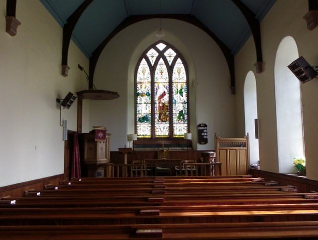 A plain church interior with dark wooden pews on a wooden floor, white walls, a blue-painted timber ceiling and a stained glass window above the pulpit.