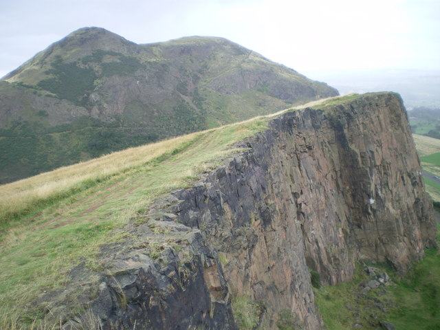 A grassy hill rising beyond the sheer edge of a basalt cliff, with green fields and a hazy horizon far below.