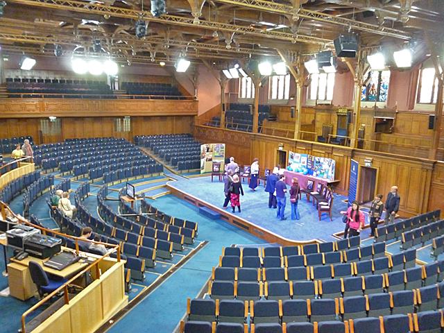 A large panelled hall with tiered blue seats curving round a central open floor, a gallery above and a timber roof.