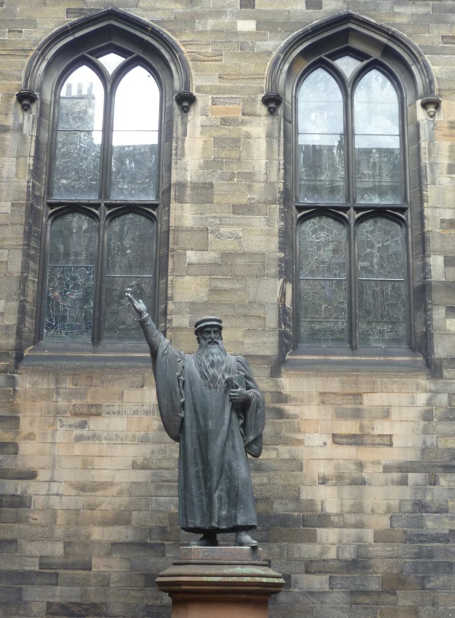 A bronze statue of a bearded man in a long gown with one arm raised, standing on a plinth against a sandstone wall with two tall Gothic windows.