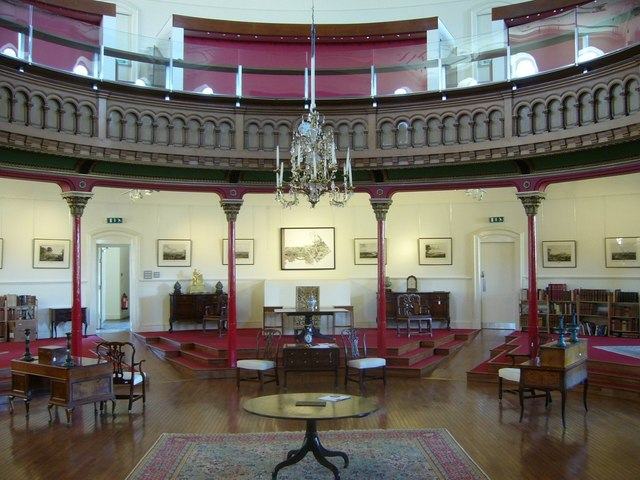 A galleried oval interior with red-painted balcony fronts, a chandelier, pictures hung along the walls and furniture set out on a wooden floor.