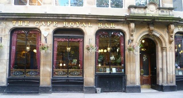 A row of tall arched pub windows with THE CAFE ROYAL BAR in gold capitals above them, hanging baskets and a dark timber door.