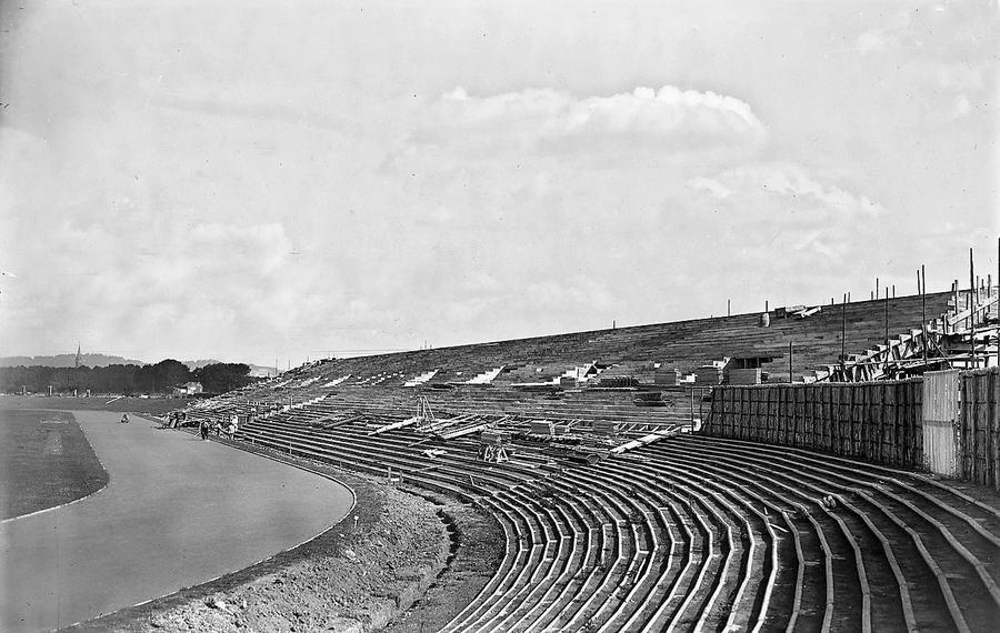 A black and white photograph of a curving open bank of terracing under construction beside a running track, with scaffolding and workmen along the top.