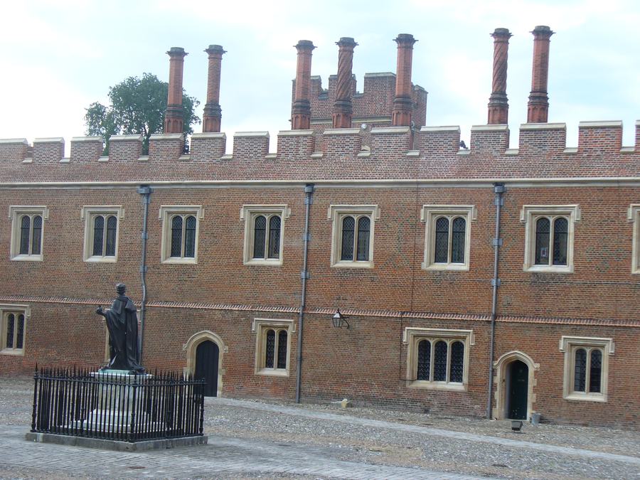 A long two-storey red brick building with battlements and a row of tall chimney stacks, seen across an empty cobbled and paved yard with a statue at its centre.