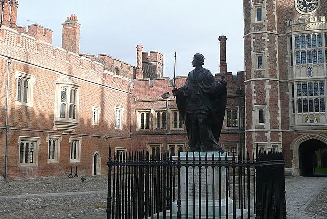 A bronze statue of a robed king inside iron railings, standing on cobbles in front of a red brick range and a tall gatehouse tower with a clock.