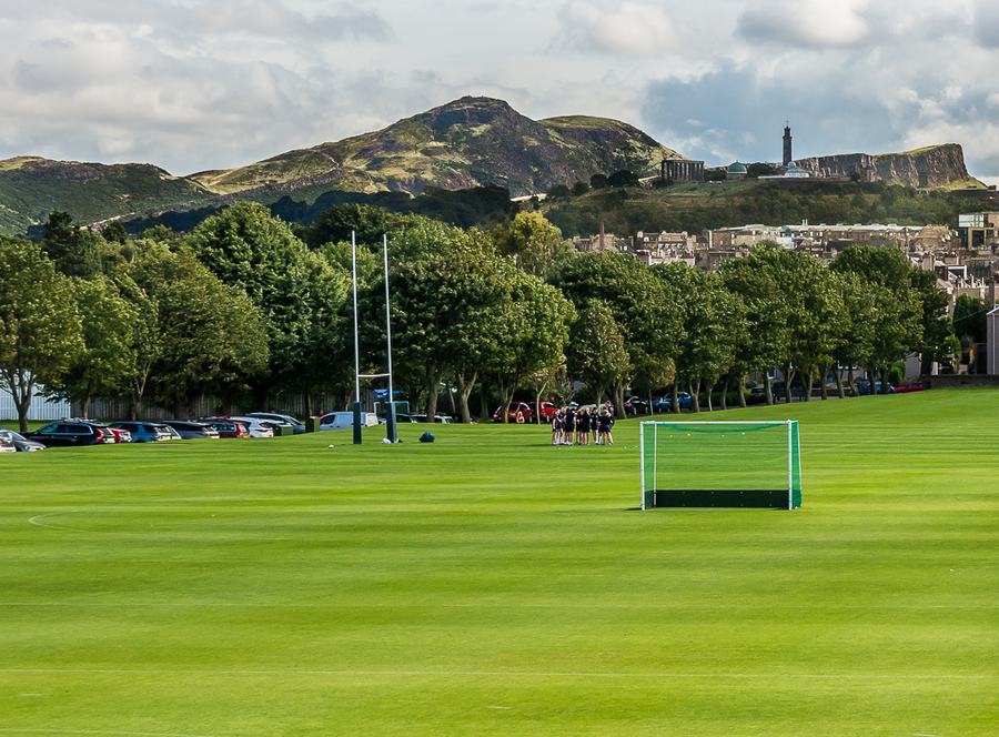 A wide flat green playing field with rugby posts and a hockey goal, a line of trees behind it and two hills on the city skyline beyond.