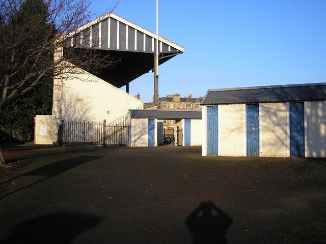 A low white timber grandstand behind a wall, with a row of turnstile huts and a tarmac approach in winter shadow.